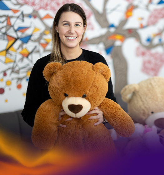 A smiling woman holds a large brown teddy bear in front of her. Orange and purple illustrated waves float beneath her.